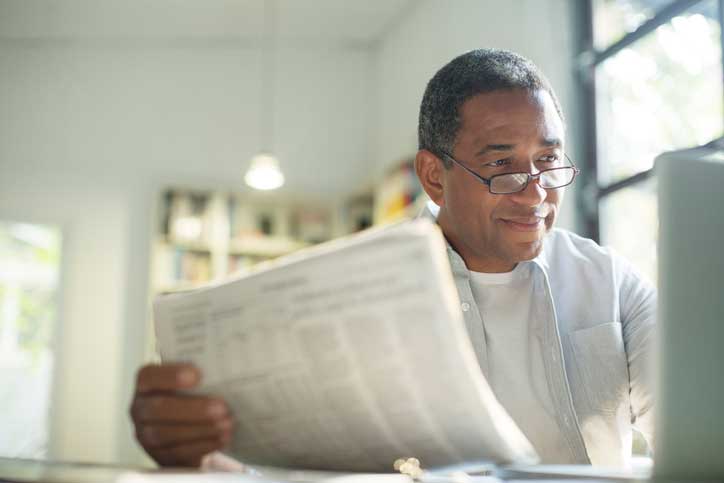 A man at his laptop while reading a newspaper.
