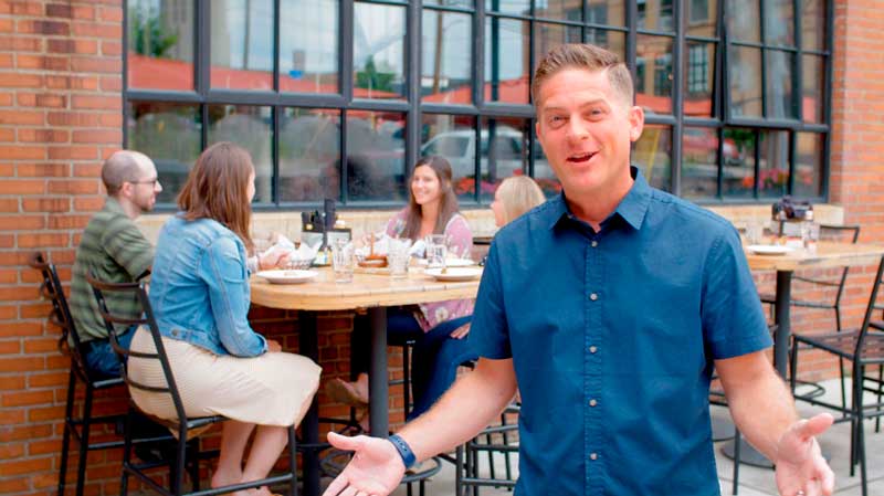 Man in blue t-shirt outside of a brewery talking excitedly in front of a group of people sitting at a table.