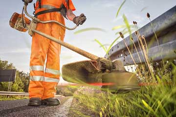 Man standing roadside using a weedwhacker to clean up the grass mobile view