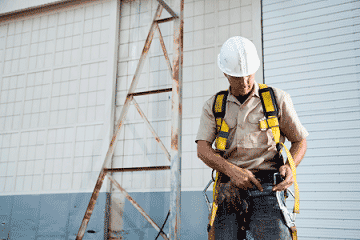 Man wearing a white hard hat standing next to ladder adjusting a fall harness